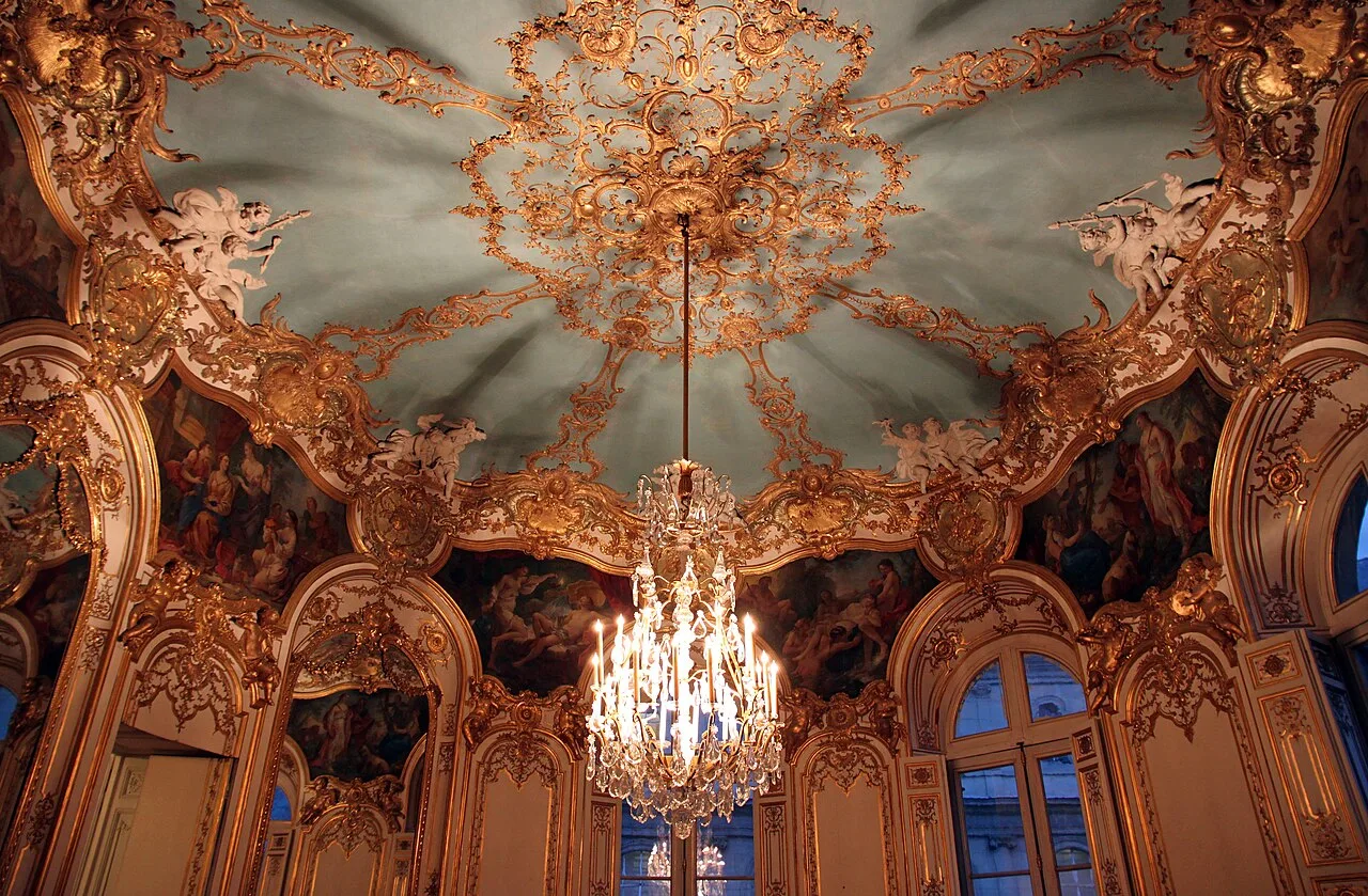 The upper white walls and gold panelling of an oval room, curving seamlessly into a domed ceiling adorned with gilded stucco vines and framed oil paintings.